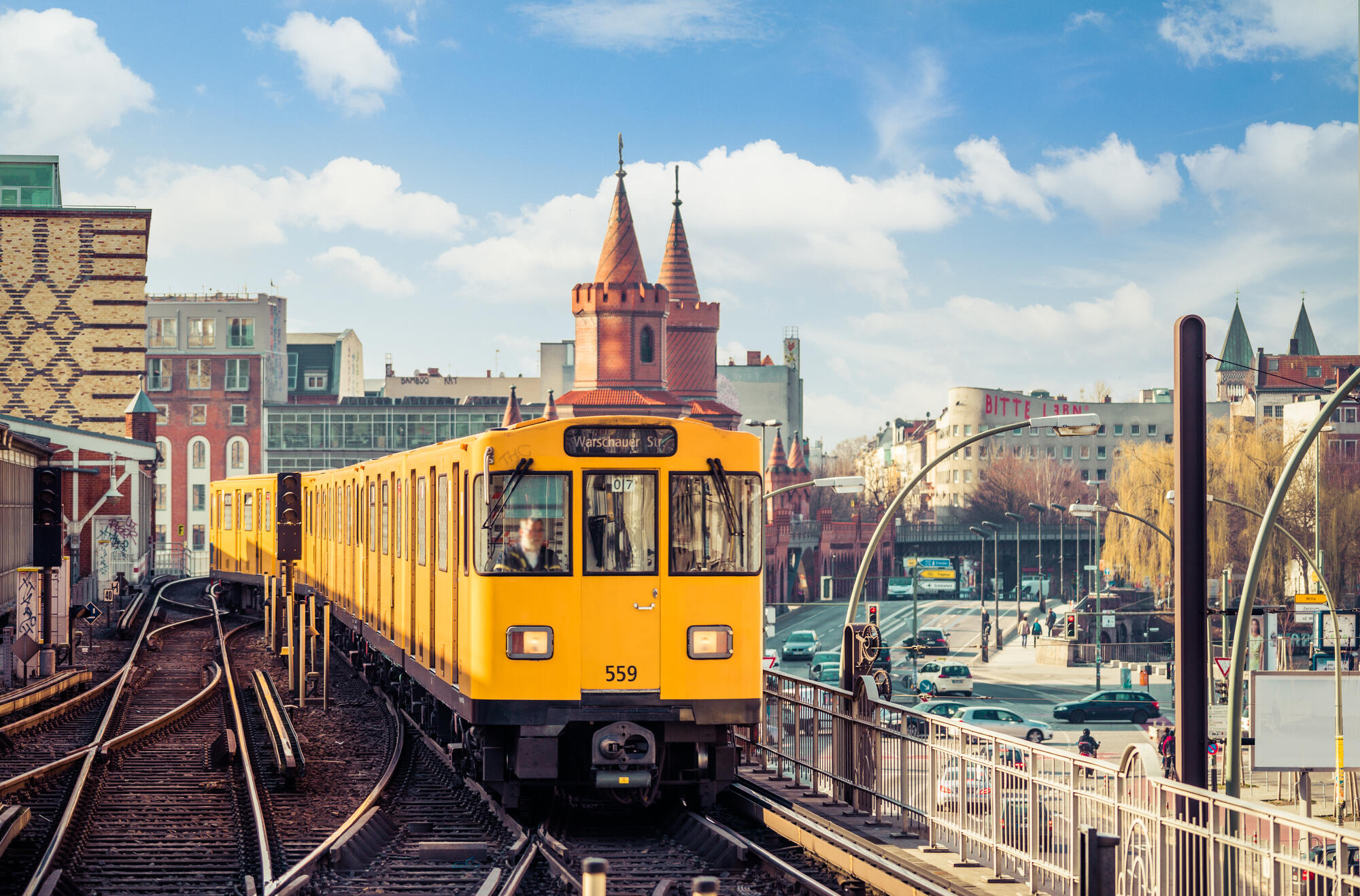 Eine gelbe U-Bahn fährt vor den roten Türmen der Oberbaumbrücke; darüber stehen helle Wolken.