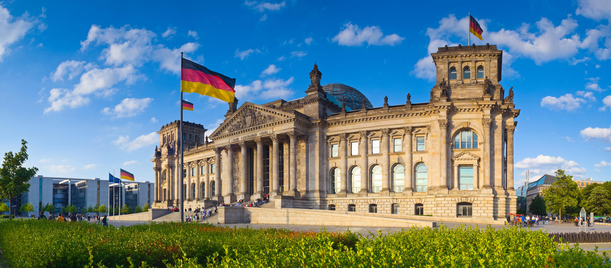 Die Westfassade des Reichstags mit wehenden deutschen Flaggen, blauem Himmel und weißen Wolken.