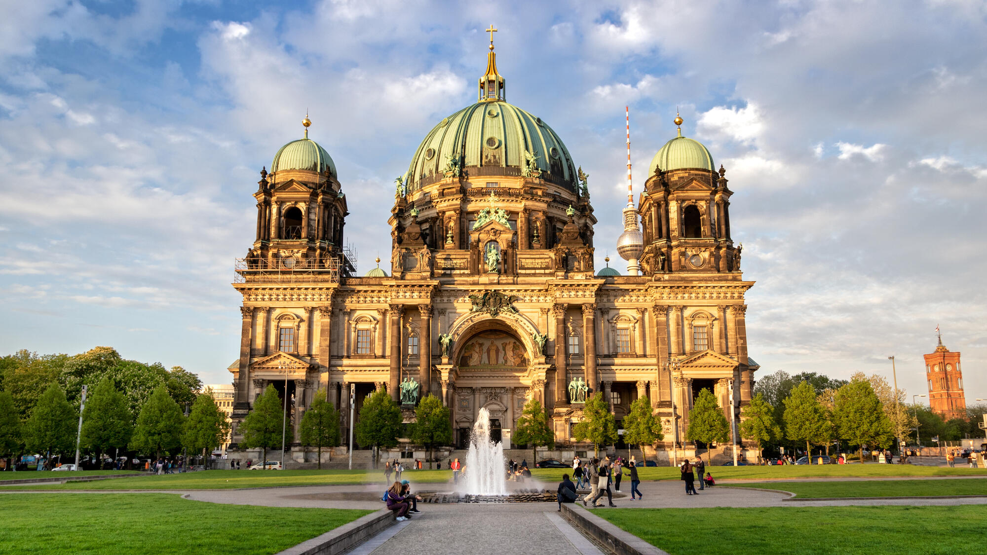 Der Berliner Dom hinter dem Springbrunnen und den Rasenflächen des Lustgartens unter einem wechselnd bewölkten Himmel.