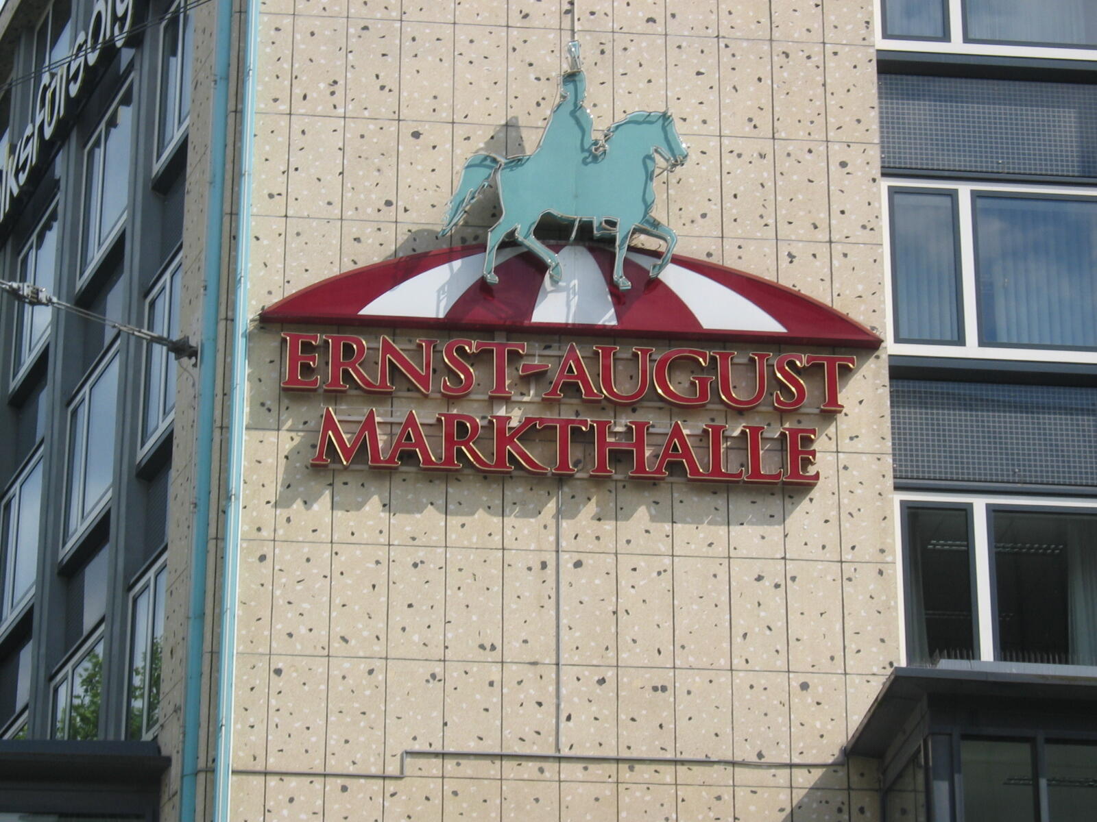 Schild mit der Aufschrift Ernst-August-Markthalle, rotem Markisendach und Pferdefigur an einer Gebäudefassade.