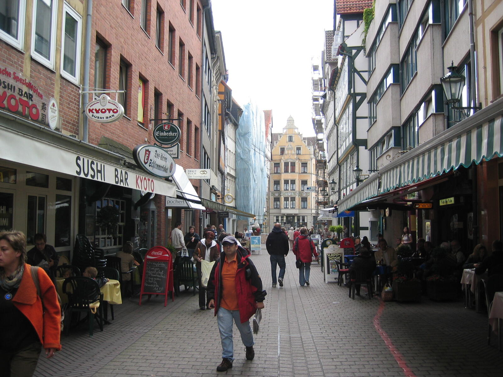 Menschen gehen durch eine schmale gepflasterte Straße mit Restaurants, Außensitzplätzen und hohen Gebäuden.