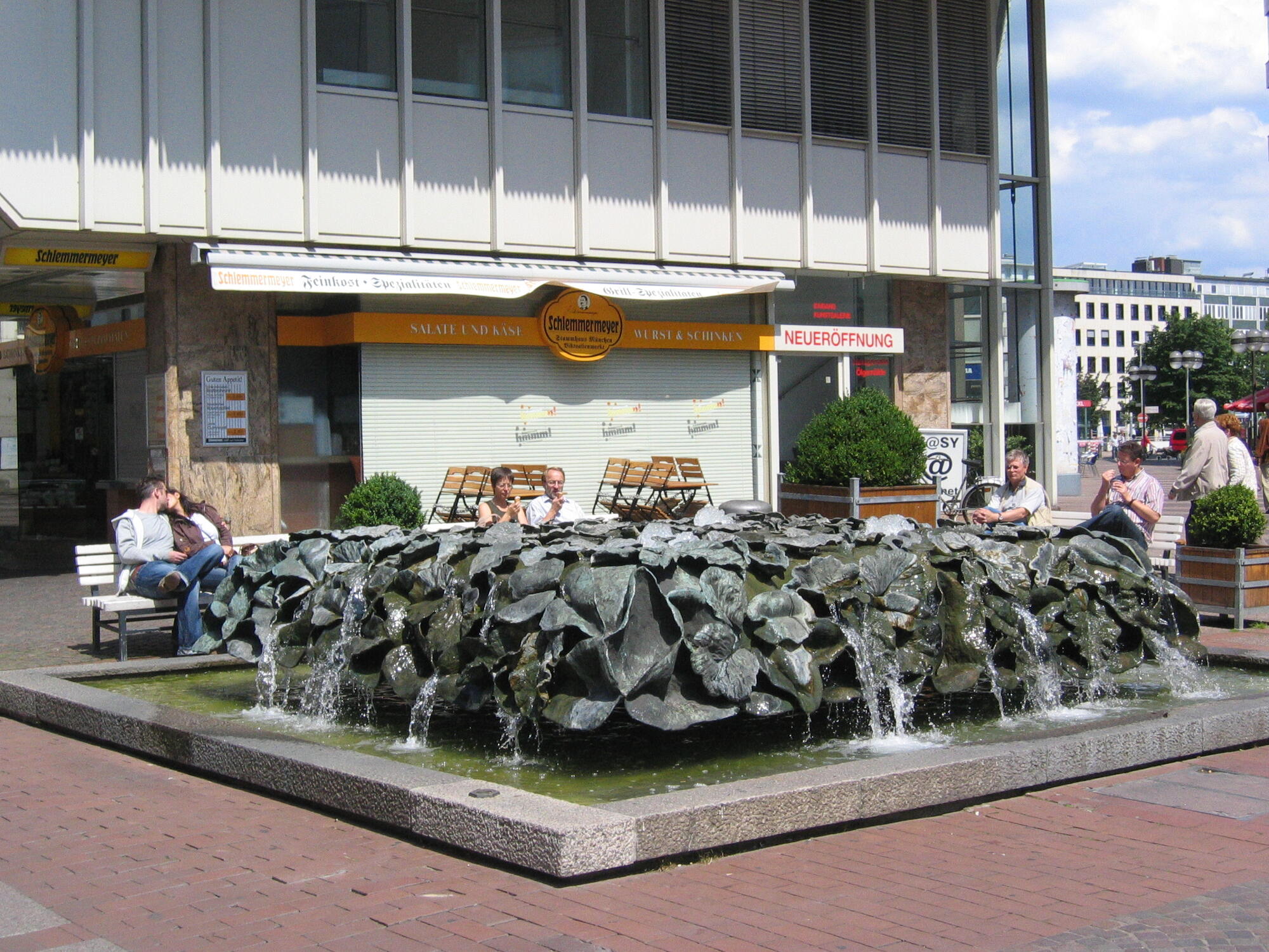 Steinbrunnen mit kleinen Wasserfällen auf einem Platz vor einem Café, umgeben von sitzenden Menschen.