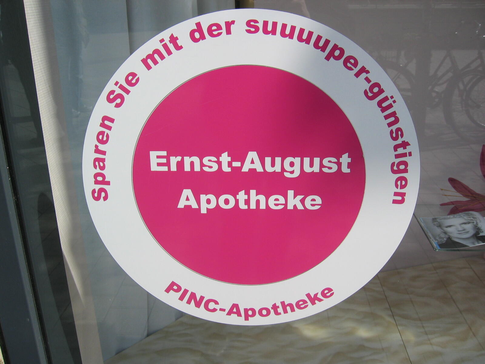Runder pink-weißer Aufkleber auf einer Glasscheibe mit Text zur Ernst-August Apotheke.