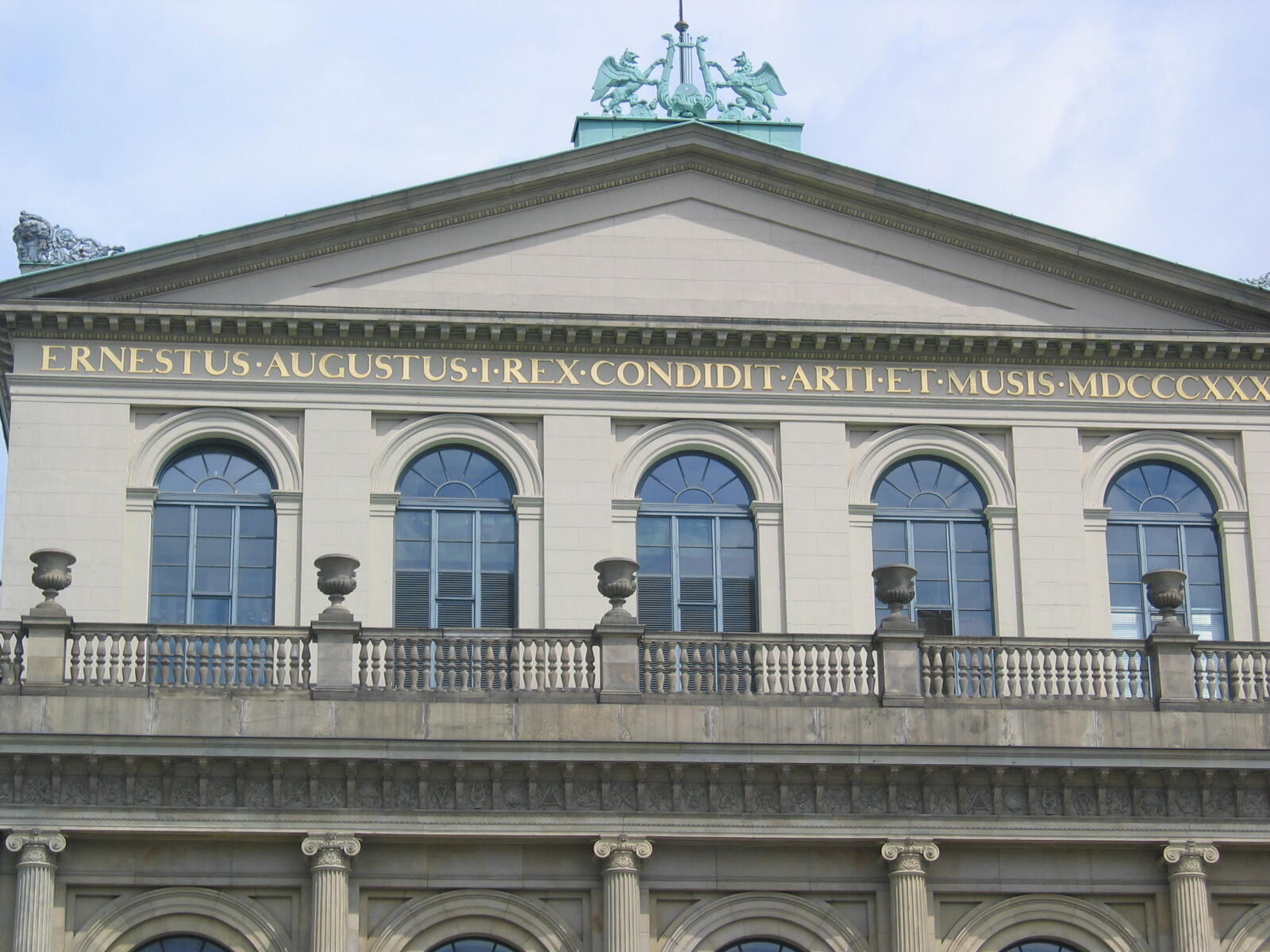 Helle klassizistische Gebäudefassade mit Dreiecksgiebel, lateinischer Inschrift, Rundbogenfenstern, Balustrade und Säulen.