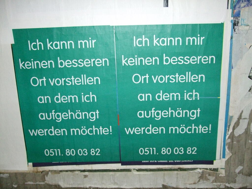 Zwei grüne Plakate mit weißem deutschem Text hängen nebeneinander an einer beschädigten Wand.