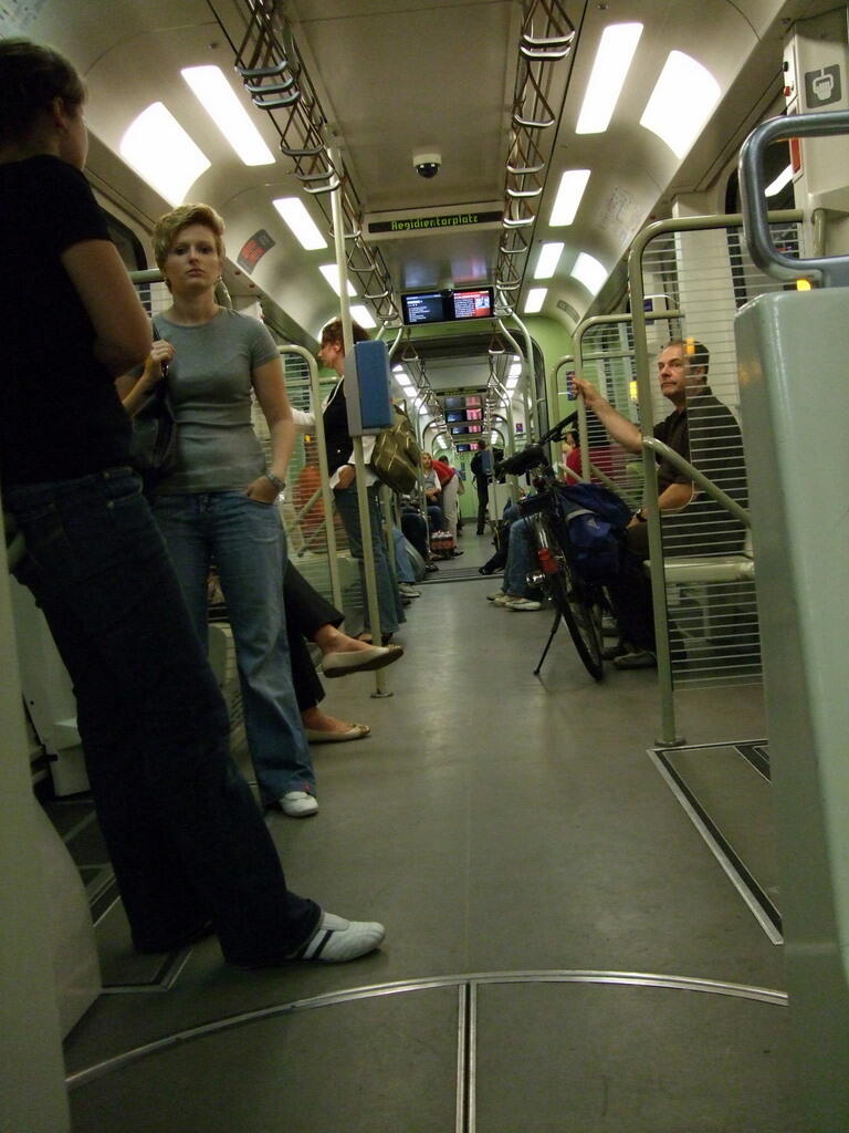Mehrere Fahrgäste stehen und sitzen im Innenraum einer hell beleuchteten Straßenbahn.