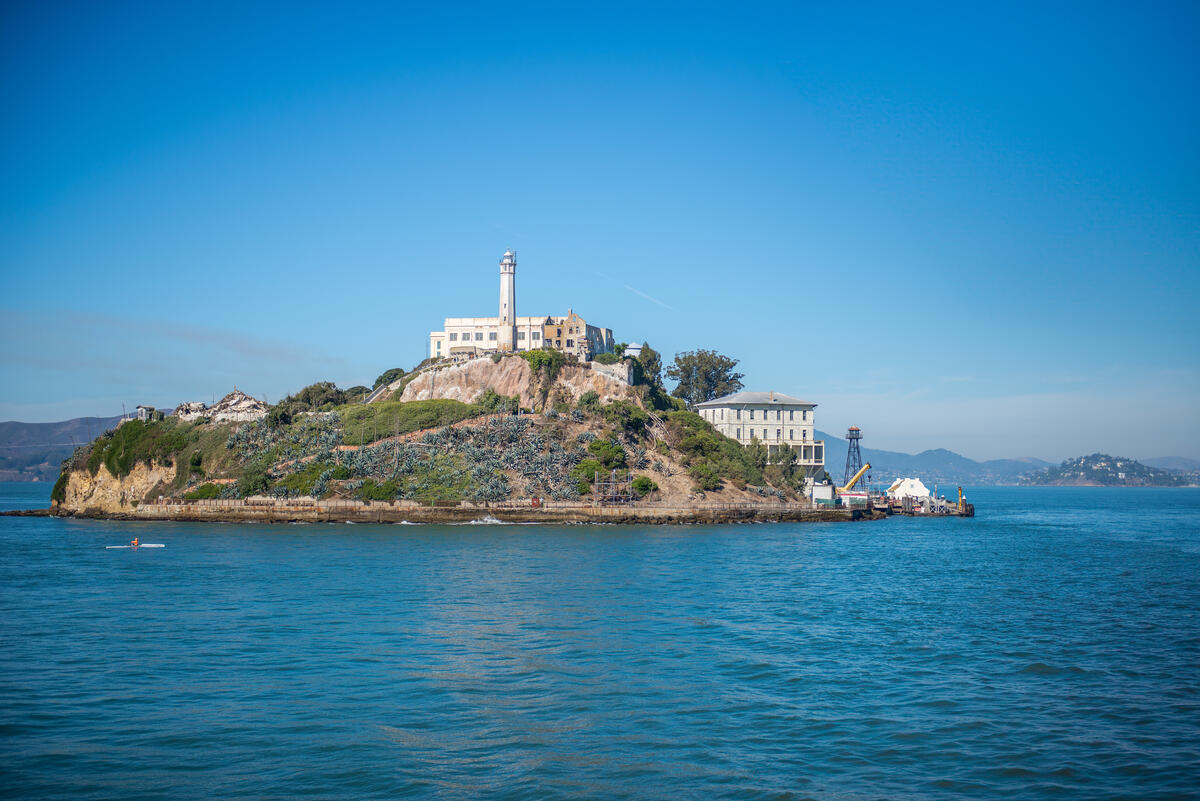 Helle Gebäude und der Leuchtturm stehen auf der felsigen, bewachsenen Insel über dem blauen Wasser der Bucht.