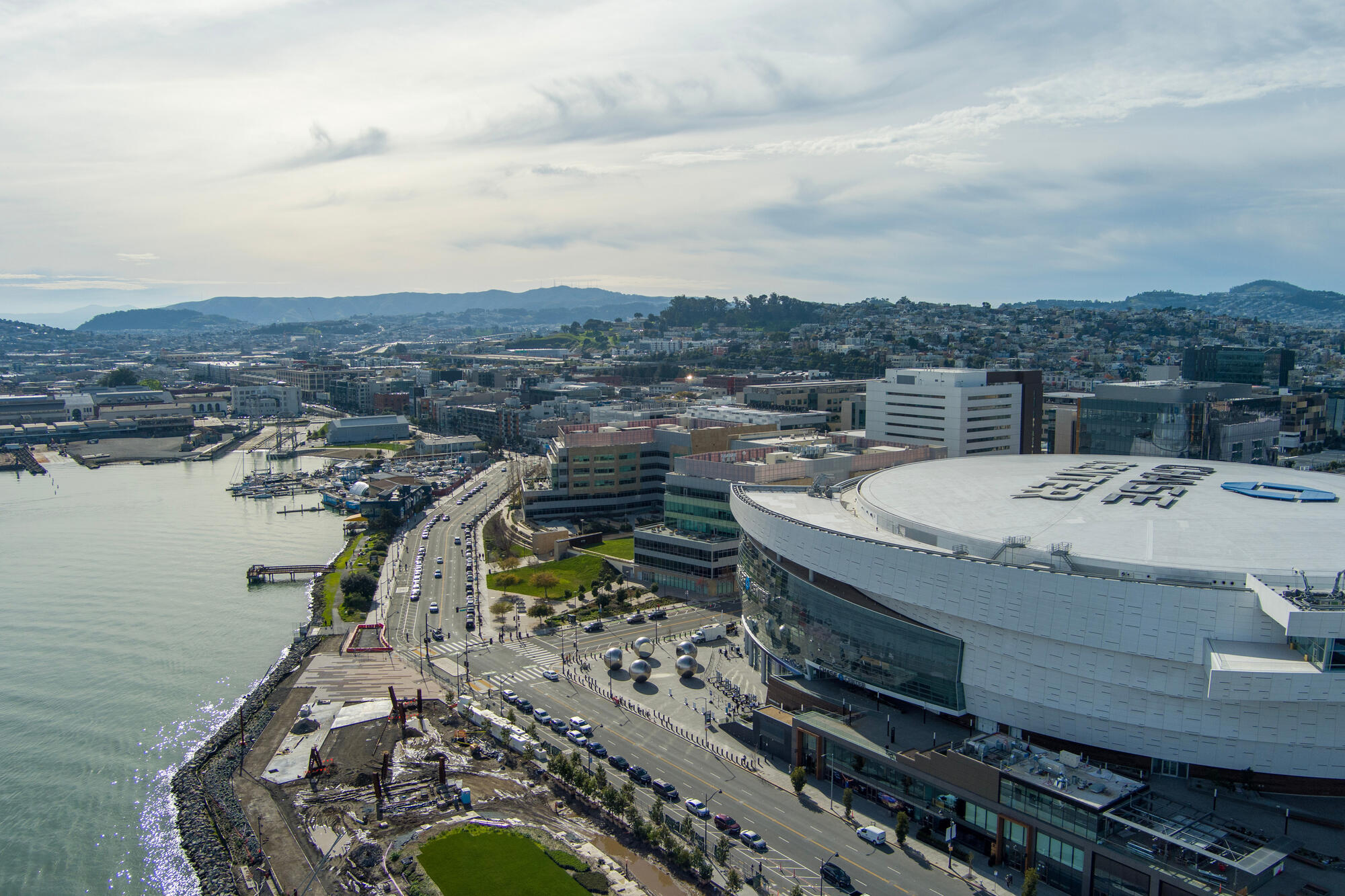 Die Luftansicht zeigt die geschwungene Arena, ihre helle Außenhaut und die Uferstraße entlang der Bucht.