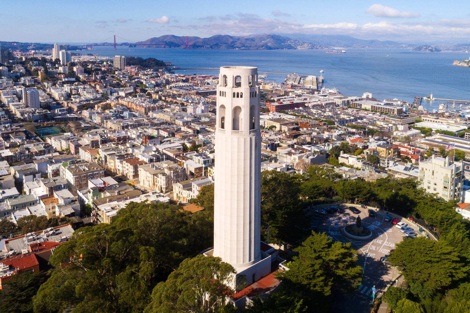 Eine erhöhte Ansicht zeigt den hellen Coit Tower zwischen Bäumen, dichten Wohnvierteln und der Bucht.