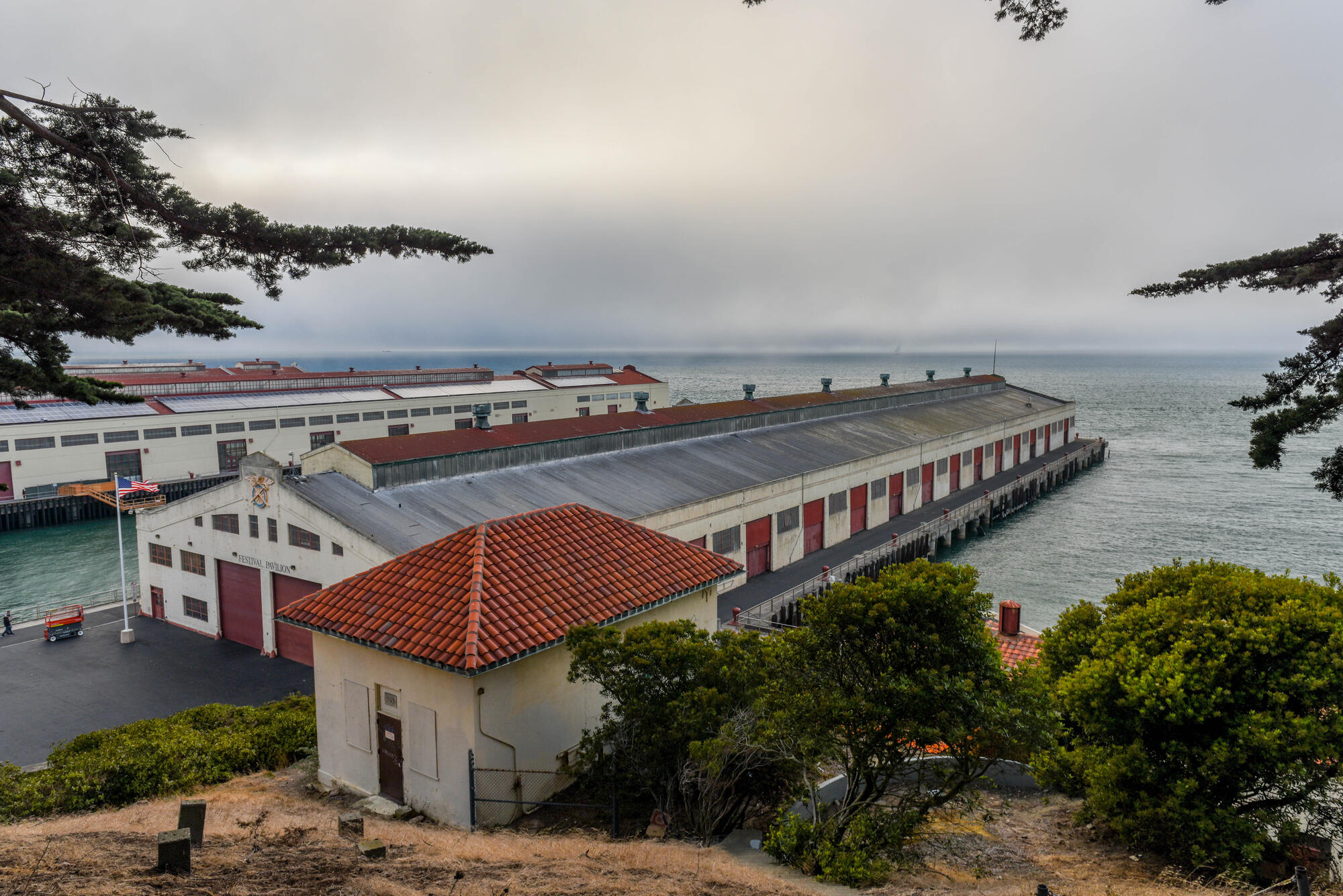 Lange Hallen mit geneigten Dächern reichen bei bedecktem Himmel in die Bucht; im Vordergrund stehen Bäume und ein kleiner Ziegelbau.