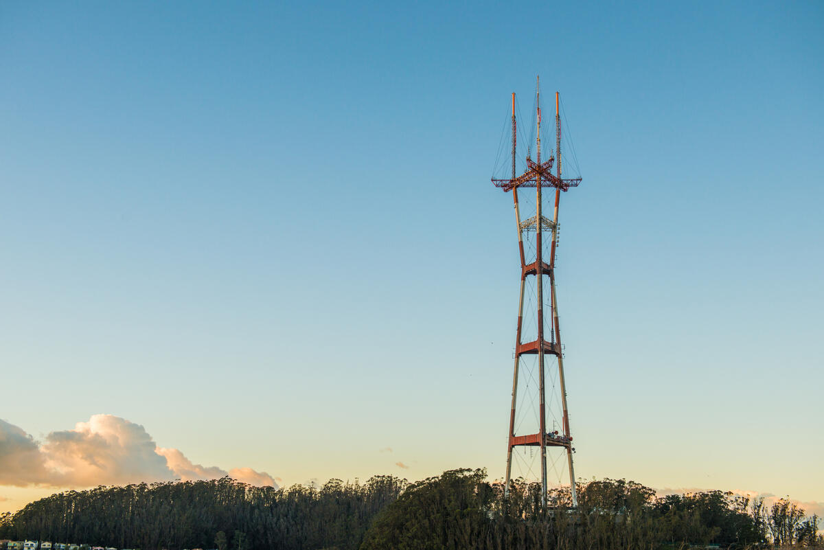 Die rot-weiße Stahlkonstruktion des Sutro Tower ragt über eine dunkle Baumlinie in den hellen Himmel.