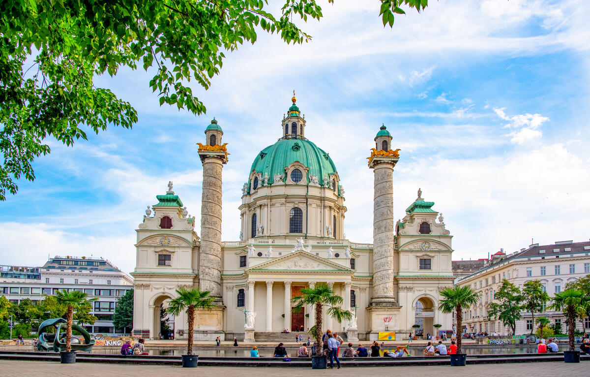 Die Karlskirche mit Kuppel und Säulen am Karlsplatz in Wien; davor sitzen Menschen am Wasserbecken.